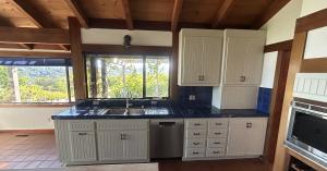 A bright residential kitchen featuring newly refinished off-white beadboard cabinets. The kitchen design includes dark blue tile countertops, a stainless steel double sink and dishwasher, natural wood ceiling beams, and large picture windows that look out