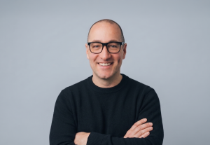 Justin Biskin, a man with a shaved head and glasses wearing a black sweater