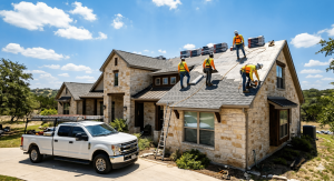 Roofing contractors installing shingles on a residential home under a clear blue sky, with a white truck parked nearby.