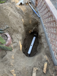 A view into a deep, freshly excavated pit revealing a section of white PVC pipe being repaired during a professional sewer repair.