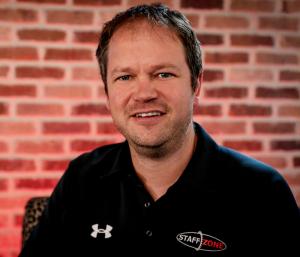 Portrait of Eric Schwartz, Staff Zone Director of Safety wearing black staff zone logoed polo shirt. White man short brown hair on red brick wall background.