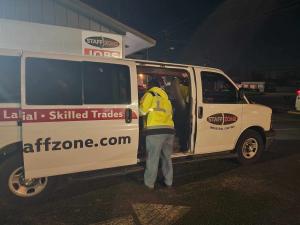 Staff Zone van driver checks that all passengers are buckled up before departure while workers wearing reflective safety gear board a company transportation van at night outside a Staff Zone branch location.
