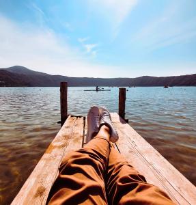 A person relaxing on a wooden dock, legs outstretched, overlooking a calm lake and mountains on a clear day.