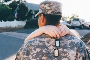 A service member in camouflage uniform and cap is embraced from behind by a loved one on a residential street at sunset.