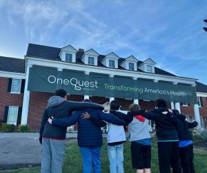Six young boys stand arm-in-arm with their backs to the camera, looking toward a large two-story brick building with white columns. A large green banner stretched across the front of the building reads, "OneQuest Health Transforming America's Health." The