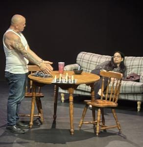 Felix stands beside a table with a chessboard while Alix sits on a couch during Scene 1 of Dominant Animals, showing a tense family conversation on stage.