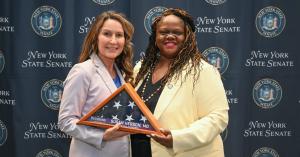 Senator Lea Webb presents Veteran Hall of Fame award to Navy Veteran Susan Herson, MD holding folded, framed American Flag
