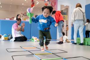 一名幼兒在 TuniTuni 故事體能課堂中,手持花朵造型教具跳過彩色地面梯。