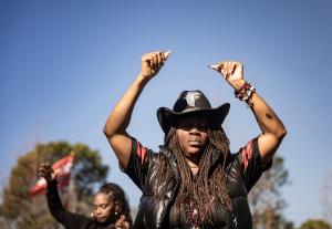 A close-up photograph of a Black woman with her eyes closed and arms raised in a moment of celebration or reflection. She is wearing a black cowboy hat with the Atlanta Falcons logo, a black puffer vest, and jewelry. In the background, another woman stand