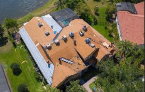 Aerial view of roof installation on a large house. Workers apply new shingles, with visible underlayment. Surrounding area includes trees and a nearby lake.
