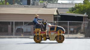 A worker in a straw hat operating heavy paving equipment to compact freshly laid asphalt.