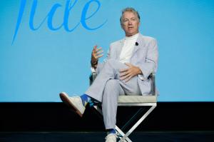 Senator Rand Paul, a middle-aged White male in a light gray suit with white pinstripes and a white turtleneck on sitting on a director's style chair. His legs are crossed and a light blue background is behind him with some words cut off.