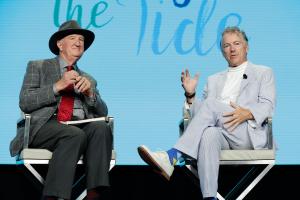 A middle-aged White male in a light gray suit with white pinstripes and a white turtleneck on sitting on a director's style chair. His legs are crossed and a light blue background is behind him with words cut off. To his left is a older white male in a dark suit. & hat.