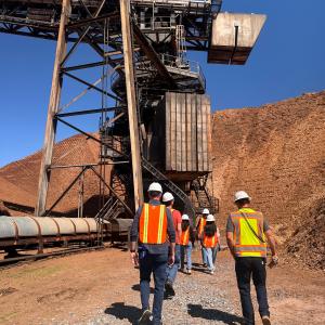 People in hard hats and safety vests walk through a wood products facility with large piles of wood fiber and industrial equipment.