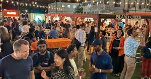 Crowd networking at an evening Boston AI Week opening party at Island Creek Raw Bar in WS Development's Seaport in Boston, with string lights and vendor stalls