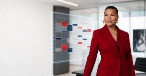 Confident Black woman executive in a deep-red blazer standing in a modern white office with a translucent holographic weekly calendar beside her. Red and navy time blocks appear on the calendar, with some faded to suggest lost hours.