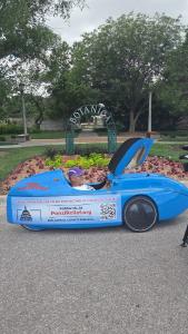 Aaron Novinger sits inside his blue velomobile, parked in front of the Botanica gardens sign in Wichita, Kansas, before pedaling toward Kansas City on his 2,000-mile charity ride for Ponzi scheme victims.