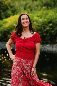 Justine Collard, praticienne périnatale et doula certifiée AQD, souriante en robe florale rouge devant un fond de verdure naturelle. Fondatrice de Naître & Être En harmonie, elle accompagne les familles de la grossesse à la naissance en Montérégie et dans