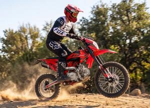 A helmeted rider in full protective gear rides a red FRP Moto FX125 125cc gas dirt bike on a dirt trail, kicking up dust during a turn.