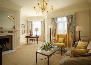 Bespoke luxury furniture inside a Savoy Hotel London suite, showcasing a gold Murano-style glass chandelier, a yellow wingback armchair, a classic mahogany writing desk, and an elegant fireplace mantelpiece available in the public auction.