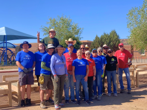 A large group of approximately 25 volunteers wearing blue and red Sleep in Heavenly Peace Arizona shirts stands together outdoors in front of completed bed frames at a school delivery site in Chinle, Arizona. The wooden beds are visible behind them, and a