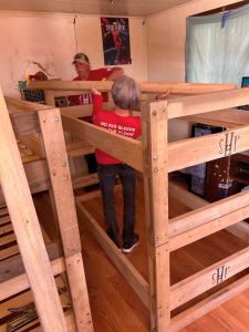 A woman in a red Sleep in Heavenly Peace Arizona shirt stands inside a wooden bunkbed frame, demonstrating its sturdy construction. The bed frame displays the "SHP" branding on its wooden headboards. The indoor setting shows additional beds stacked in the