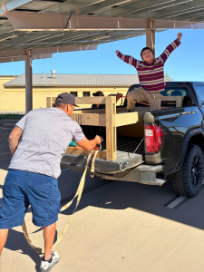 Volunteers load wooden bed frames onto a black pickup truck under a metal carport structure. In the foreground, a man in a gray shirt secures a bed frame with rope. In the truck bed, a young child in a burgundy striped shirt raises both arms in celebratio