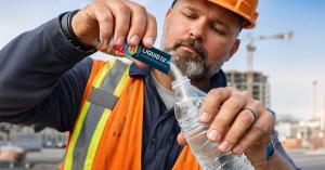 A construction worker in an orange safety vest and hard hat pouring a Liquid I.V. Hydration Multiplier packet into a water bottle on an active job site.