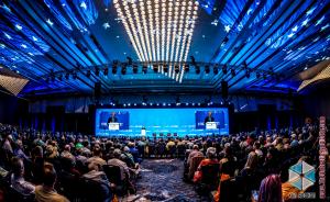 A wide-angle view of the lights, stage, and audience at FreedomFest in Las Vegas. Rows of people seated from behind, colorful blue and white lights artistically linke the ceiling in a convex pattern.