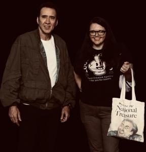 A smiling woman wearing glasses and a black graphic t-shirt poses next to a man in a dark olive bomber jacket and white t-shirt, against a dark background. She holds a cream-colored tote bag printed with the words 'You're my National Treasure' and a face