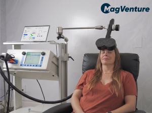 Patient sitting in chair with TMS coil over her forehead. The MagVenture TMS system is next to her as she receives PTSD treatment using MeRT.