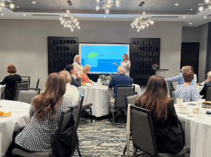 A woman stands at the front of a room full of people sitting at tables while she delivers opening remarks at The Parmenter Foundation's annual grantee breakfast.