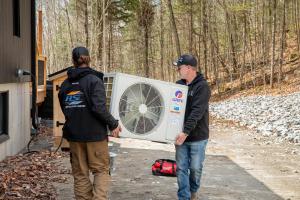 Deux techniciens d'Air Solution Confort transportent ensemble, à bout de bras, une unité extérieure de thermopompe murale GREE Inverter fonctionnant au réfrigérant écologique A2L R32, jusqu'à son point d'installation final près d'un chalet