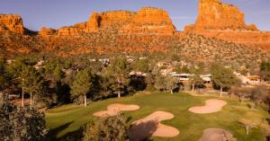 Aerial view of Oakcreek Country Club's green fairways and sand bunkers surrounded by lush trees, with the towering orange and red sandstone buttes of Sedona, Arizona rising in the background at golden hour.