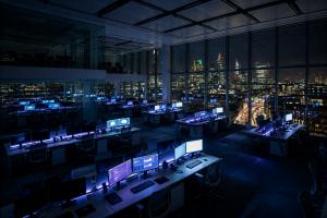A dimly lit office at night, empty desks and glowing screens visible through floor-to-ceiling windows with a city skyline in the background.
