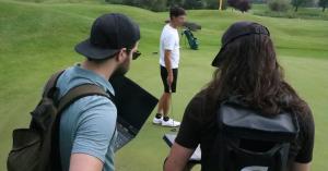 Three people on a golf green: a golfer in a white shirt holding a putter in the background (face pixelated for privacy), and two PsyLinks team members in the foreground, one holding an open laptop, recording data outdoors.