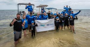 AquaOmega and 4ocean team members standing in shallow ocean water holding an AquaOmega x 4ocean banner during a coastal cleanup initiative.