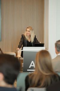A woman in a black blazer speaking from a podium with an AI Insiders sign to a seated audience.