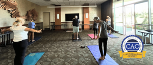 A group of adults practicing yoga on mats in a well-lit room with large windows and a "Certified Autism Center" badge overlay.