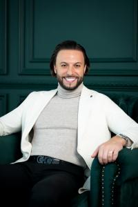 Maher Elusini seated against a dark green background, wearing a white jacket and black shirt.