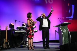 A man and a woman on stage with a man making a military style salute with one hand and a statue raised up in the other hand, as the woman stares at him. On stage for Anthem Film Festival. Pretty pink and purple lights are on a screen behind them. The man is in a tuxedo.