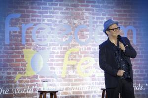 A middle-aged white male in glasses and a 40's style hat, in dark clothes against a screen that looks like a painted brick wall with the word Freedom on it, for the Punching Up comedy festival at FreedomFest.