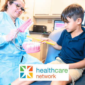 Dental professional teaching a child how to brush teeth using a large model of teeth and a giant toothbrush in a clinic.