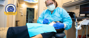 Dentist in protective gear examining patient’s teeth in a dental clinic with Certified Autism Center (CAC) logo visible.