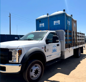 A white Barrios Site Services flatbed truck carrying blue portable toilets for delivery to a Central Valley job site.