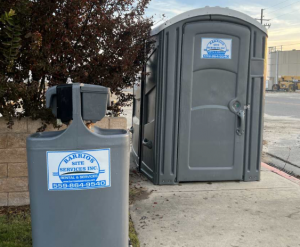 A gray Barrios Site Services portable toilet and matching handwash station situated at a job site.