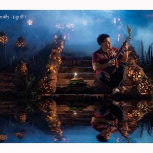 Master Vee seated beside reflective water at night, playing a traditional Phin among illuminated lanterns in a dramatic cultural performance setting.