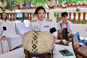 Master Vee seated at a Thai community ceremony, holding a traditional woven percussion instrument, with students and traditional musical instruments in the background.