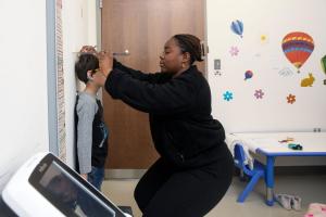 A child stands against a wall while an adult uses a flat measuring tool to check the child’s height. The scene takes place in a brightly decorated clinic exam room, with colorful wall decals of hot air balloons and flowers, a small table with crayons, and