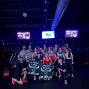 A group of people posing inside The Junk Yard Fitness, with two members holding signs celebrating their first and hundredth classes.